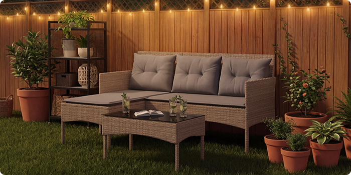 Rattanen tuinloungeset met grijze kussens, inclusief een driezitsbank, een voetenbankje en een salontafel op een gazon met snoerverlichting en potplanten