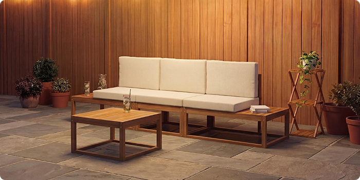 Driezits modulaire buitenbank met crèmekleurige kussens en een bijpassende lage salontafel op een stenen terras voor een warme houten muur