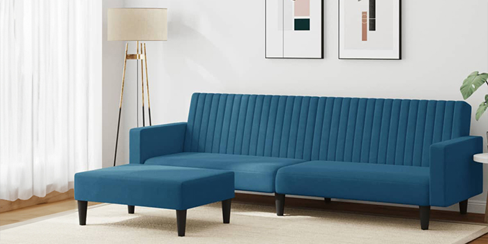 Een moderne blauwe fluwelen hoekbank met verticale stiknaden en een bijpassende poef. De bank heeft zwarte houten poten en staat in een lichte woonkamer met ingelijste kunst aan de muur en een vloerlamp in de hoek.